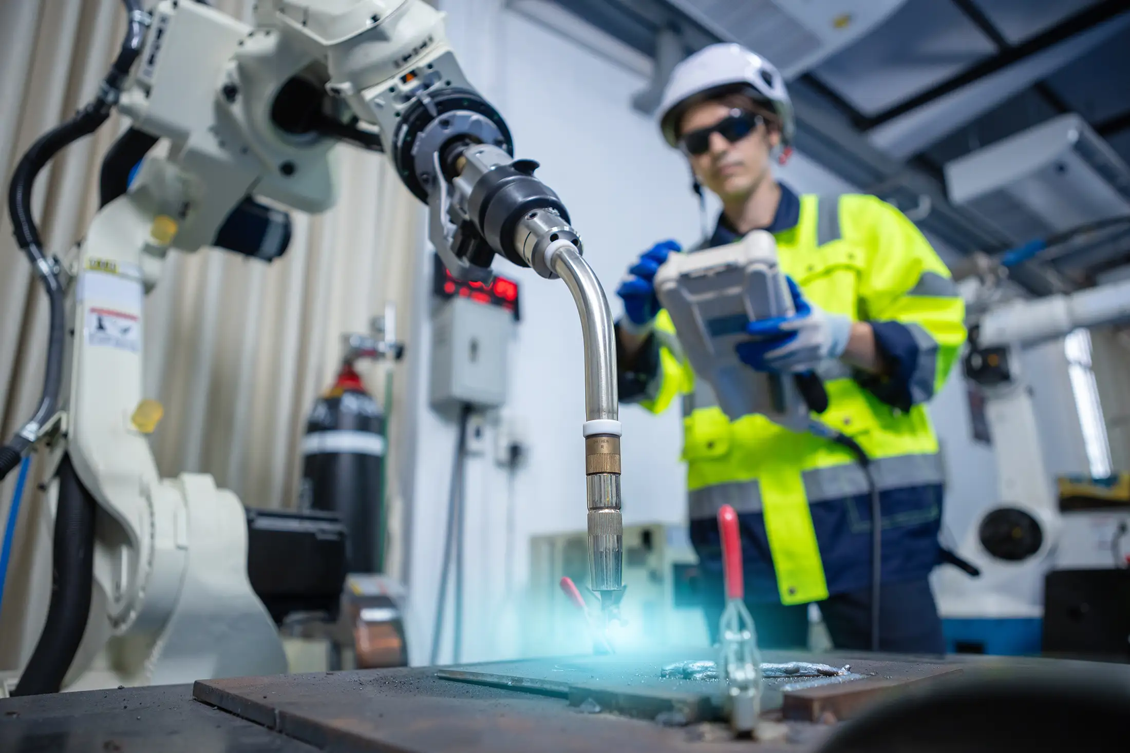 Engineer operating a robotic welding system in an industrial lab, wearing safety gear and controlling the process with a handheld device under bright blue arc light.