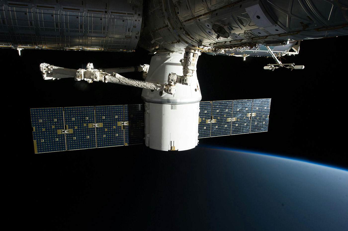 Spacecraft docked to a space station with extended solar panels, orbiting above Earth’s atmosphere with the planet’s curved horizon visible against the blackness of space.
