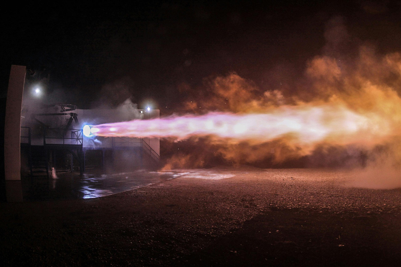 Rocket engine firing during a ground test at night, producing a bright, powerful exhaust plume of orange and purple flames with smoke and vapor illuminated by the thrust.